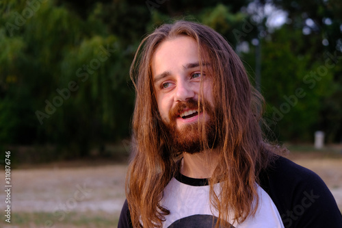 Handsome smiling young man with long hair posing at sand beach near forest. Happiness concept