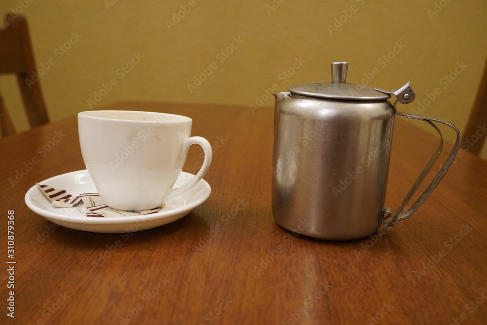 Old dirty used aluminum tea pot kettle with dirty used white tea mug. Antique Aluminum Kettle with tea cup and empty used sugar sachets. Dirty coffee and tea cup half empty on saucer on table. Old dirty used aluminum tea pot kettle with dirty used white tea mug. Antique Aluminum Kettle with tea cup and empty used sugar sachets. Dirty coffee and tea cup half empty on saucer on table.