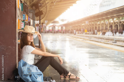 Beautiful Asian woman in a white T-shirt hidding face under straw hat from sunlight, She is sitting on floor learning a brown pillar with a black fan at train station