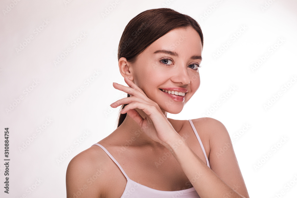 Attractive beautiful positive young woman posing alone on camera. Touch side of face with fingers and smile. Tenderness and pure. Cosmetology and dermatology. Isolated over light background. Attractive beautiful positive young woman posing alone on camera. Touch side of face with fingers and smile. Tenderness and pure. Cosmetology and dermatology. Isolated over light background.