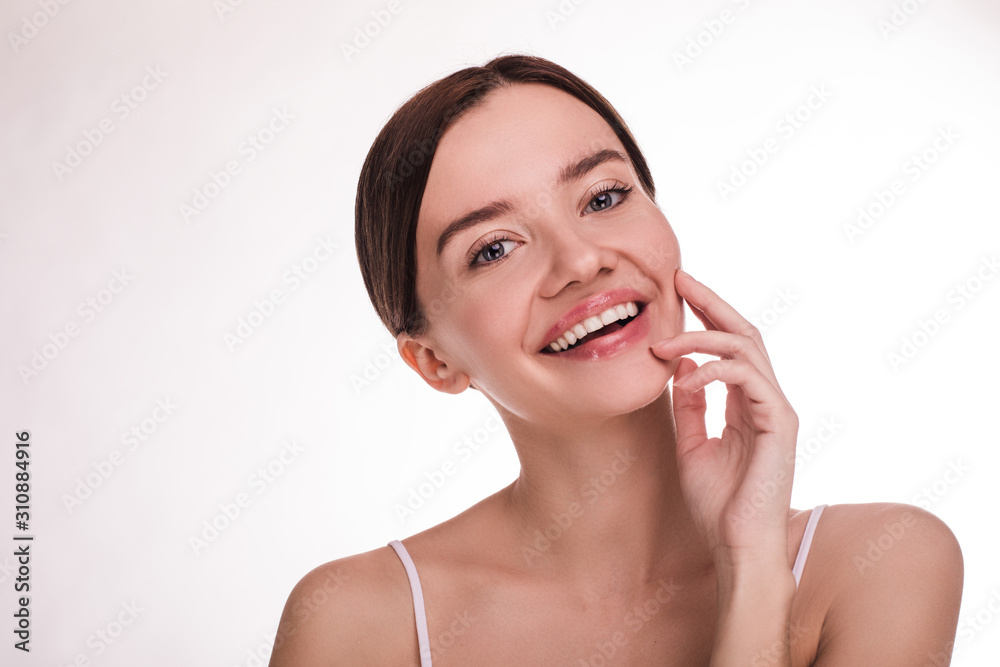 Cheerful attractive young woman posing on camera and smile. Hold hand and fingers close to face. Clear pure face skin. Cosmetology and aesthetics. Isolated over light background.