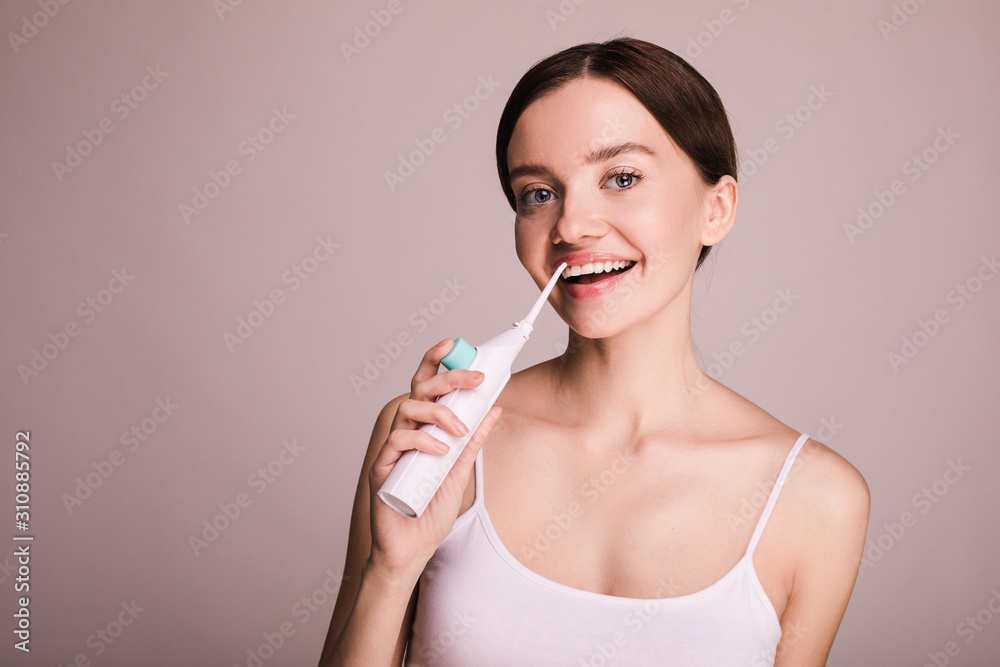 Cheerful happy young woman hold oral water irrigator and use it for interdental space. Take care about teeth. Young model stand and pose. Happy cheerful device user. Isolated over beige background. Cheerful happy young woman hold oral water irrigator and use it for interdental space. Take care about teeth. Young model stand and pose. Happy cheerful device user. Isolated over beige background.