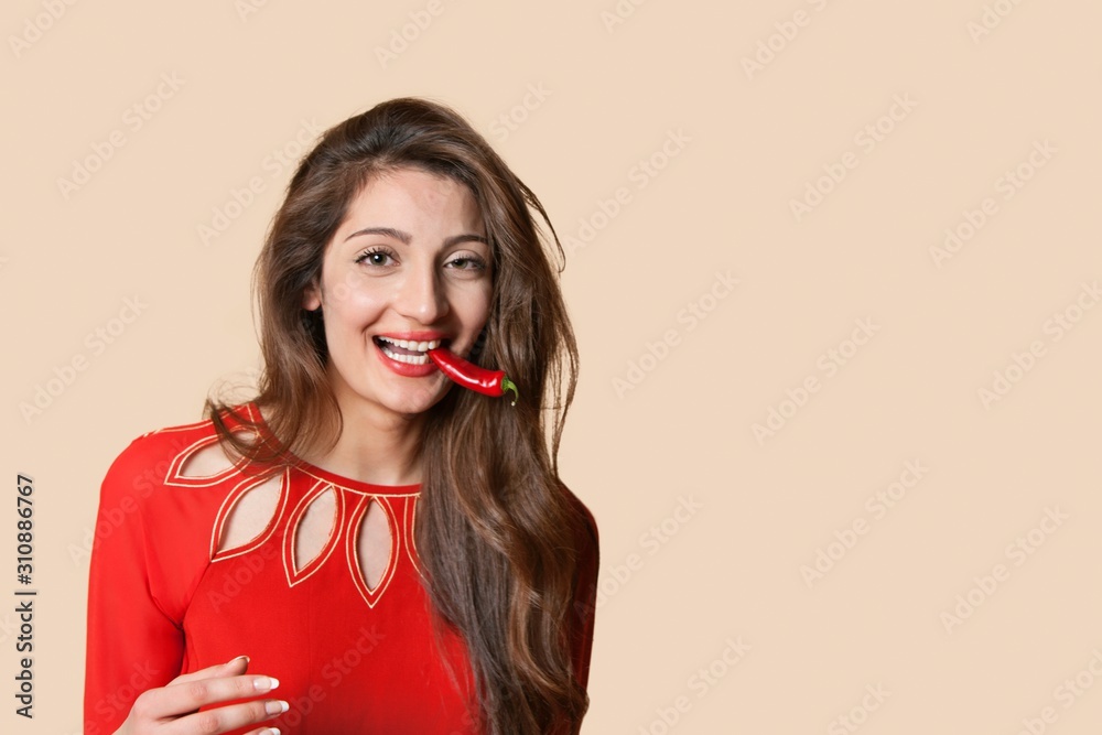 Portrait of a beautiful young woman with red chili pepper in mouth over colored background Portrait of a beautiful young woman with red chili pepper in mouth over colored background