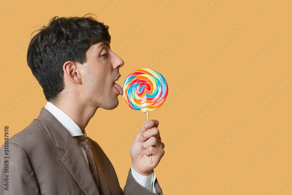 Side view of young businessman licking lollipop over colored background Side view of young businessman licking lollipop over colored background