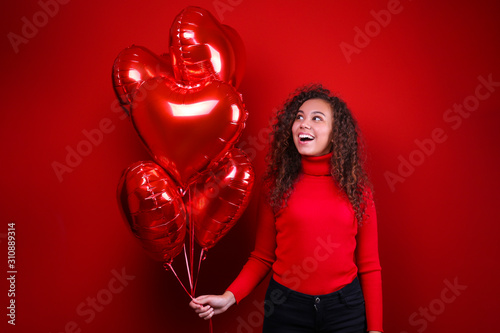 Studio portrait of young woman with dark skin and long curly hair wearing knitted turtle neck sweater over the festive red wall with heart shaped balloon. Close up, isolated background, copy space. © Evrymmnt