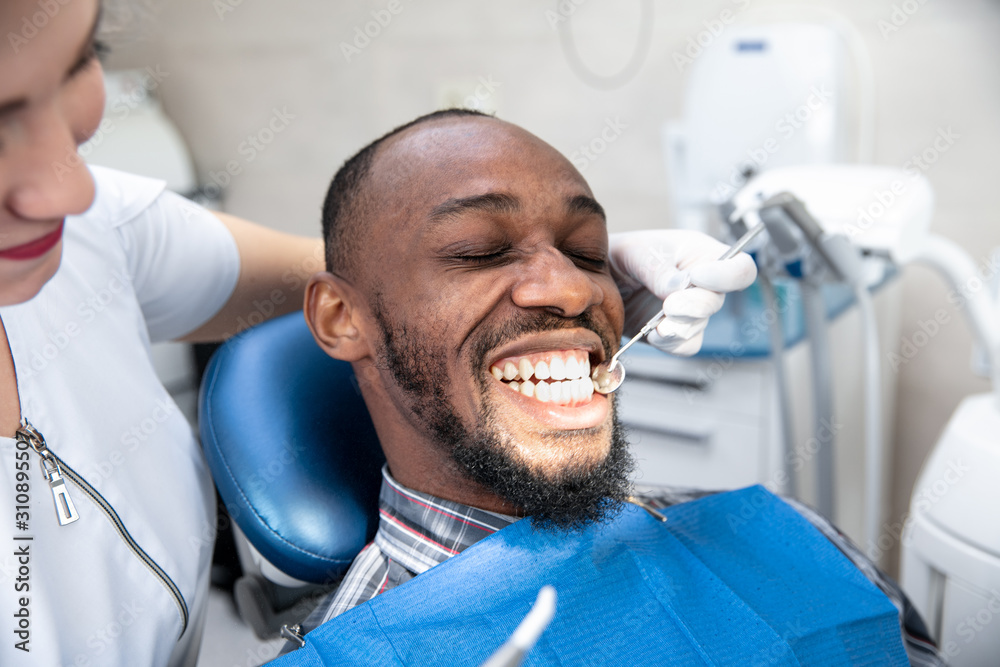 Fototapeta premium Young african-american man visiting dentist's office for prevention and treatment of the oral cavity. Man and woman doctor while checkup teeth. Healthy lifestyle, healthcare and medicine concept.