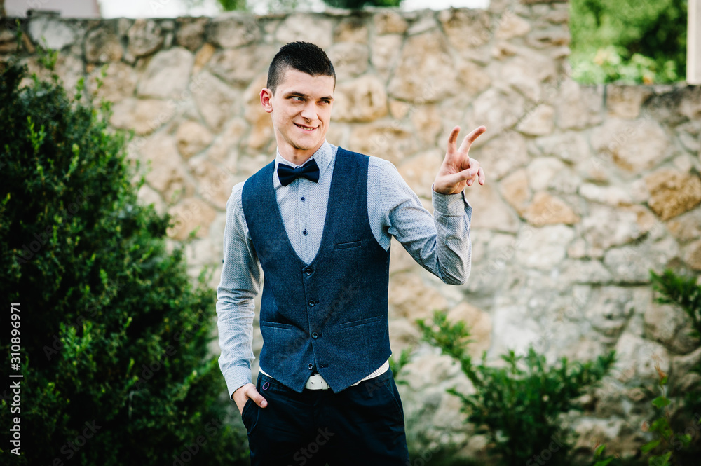 Stylish happy smile groom poses on the grass, on background in the courtyard of house. Close up. Nature. Against the background of the stone wall. A man with a butterfly. Shows two fingers. Stylish happy smile groom poses on the grass, on background in the courtyard of house. Close up. Nature. Against the background of the stone wall. A man with a butterfly. Shows two fingers.