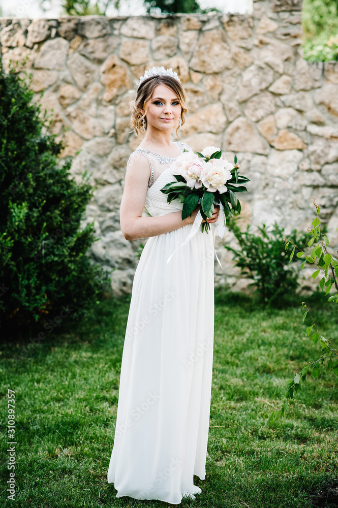 Stylish happy smile bride with bouquet of peonies with crown, stand, poses on grass on background in courtyard. Close up. Nature. Against the background of the stone wall. Looking forward.