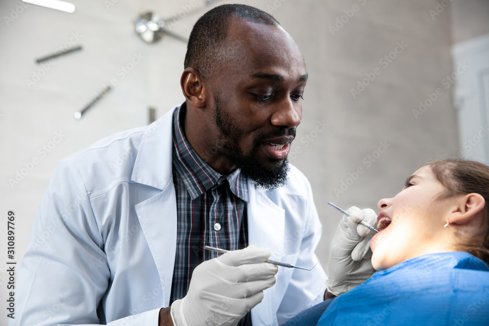 Young caucasian girl visiting dentist's office for prevention and treatment of the oral cavity. Child and african-american doctor while checkup teeth. Healthy lifestyle, healthcare and medicine Young caucasian girl visiting dentist's office for prevention and treatment of the oral cavity. Child and african-american doctor while checkup teeth. Healthy lifestyle, healthcare and medicine