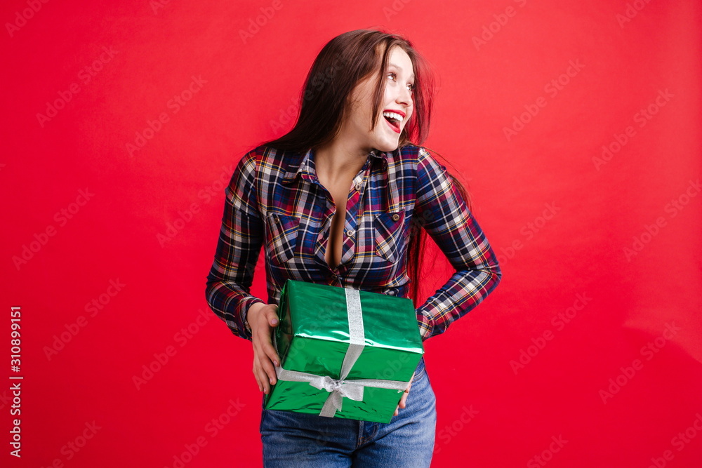 Happy girl in a plaid shirt, holding a gift box standing on a red background looking away