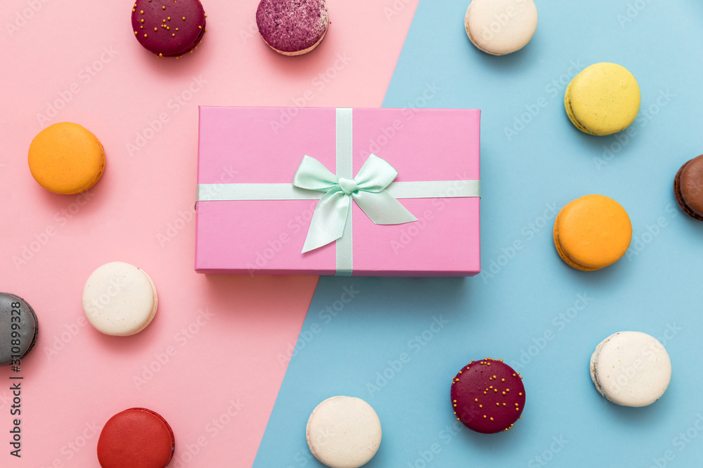 Fototapeta premium Multi-colored macaroon cakes on a pink and blue pastel background with a gift box flat lay