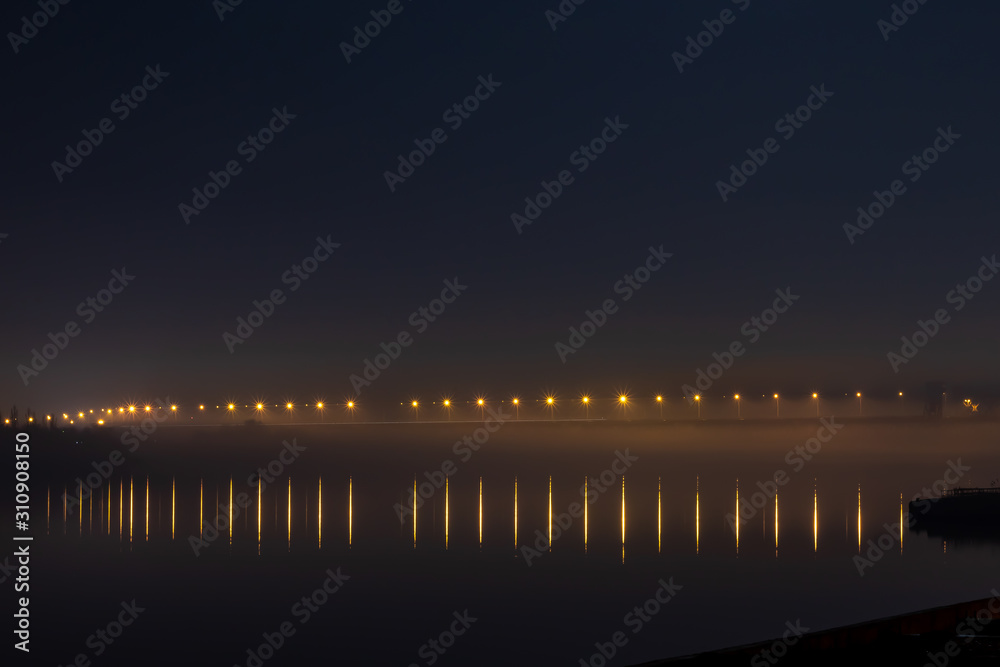 A bridge across the river lit by yellow lanterns in the fog and reflections of light in the water.