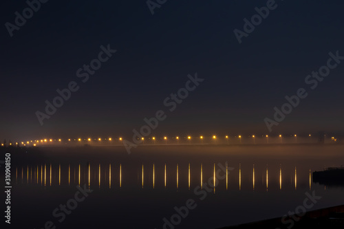 A bridge across the river lit by yellow lanterns in the fog and reflections of light in the water.