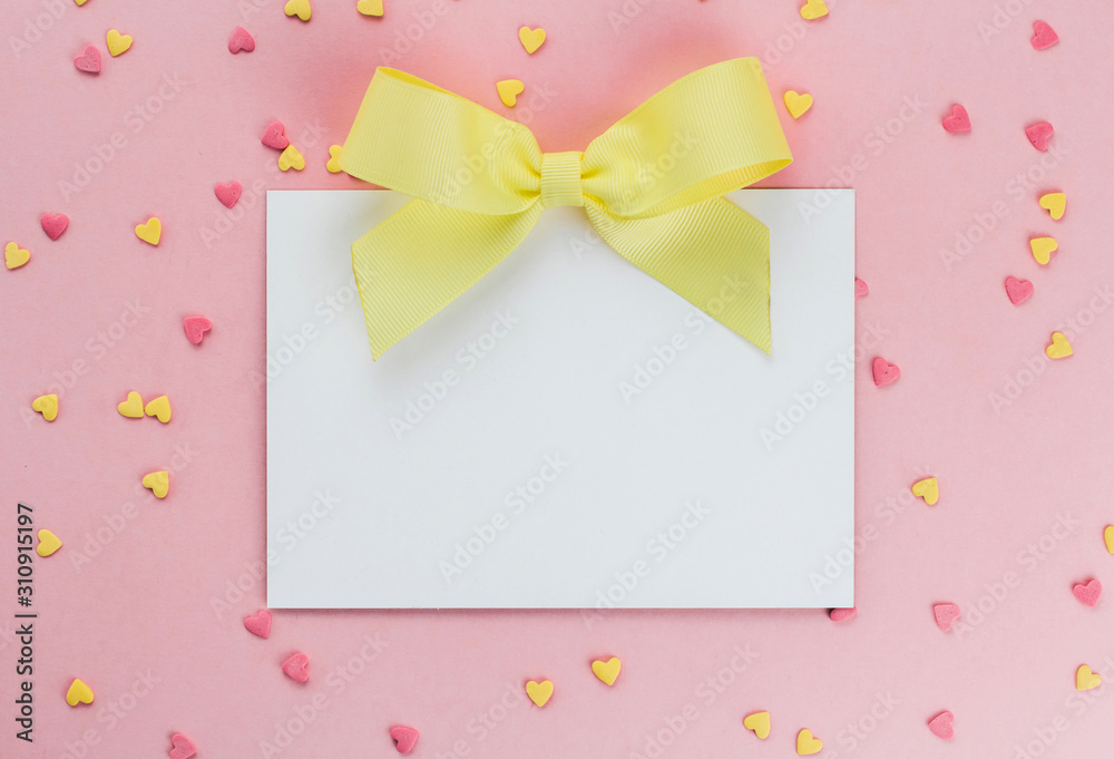  card with a yellow bow on a background of heart-shaped confectionery confetti on a pink background copy space.