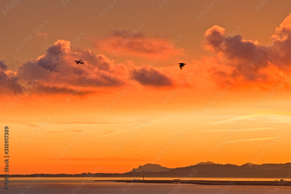 Silhouette of an airplane taking off from Nice airport, during a fiery red and orange winter sunset