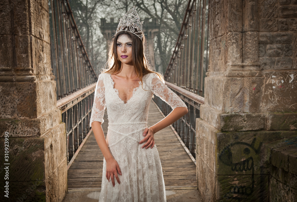 Obraz premium Lovely young lady wearing elegant white dress enjoying the beams of celestial light and snowflakes falling on her face. Pretty brunette girl in long wedding dress posing on a bridge in winter scenery