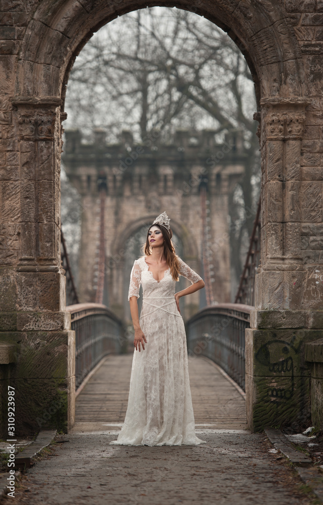 Obraz premium Lovely young lady wearing elegant white dress enjoying the beams of celestial light and snowflakes falling on her face. Pretty brunette girl in long wedding dress posing on a bridge in winter scenery