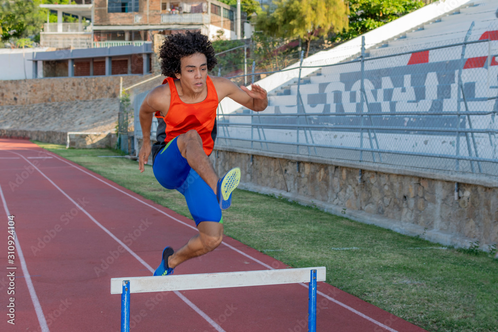 Obraz premium Latin fit young man with afro style hair in orange tank top at the running track jumping the hurdless fence on a sunny day.