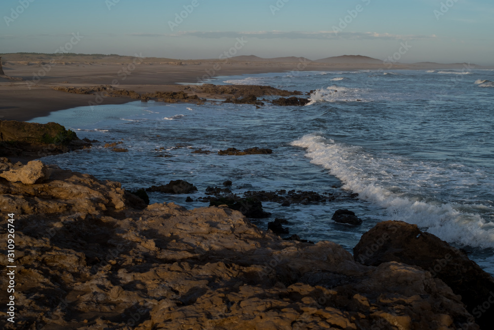 Landscape of stone bay with strong waves on sandy beach and dunes, at sunset