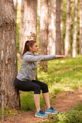Beautiful caucasian brunette in sportswear and with ponytail leaning against the tree and squatting. Fitness in nature concept.