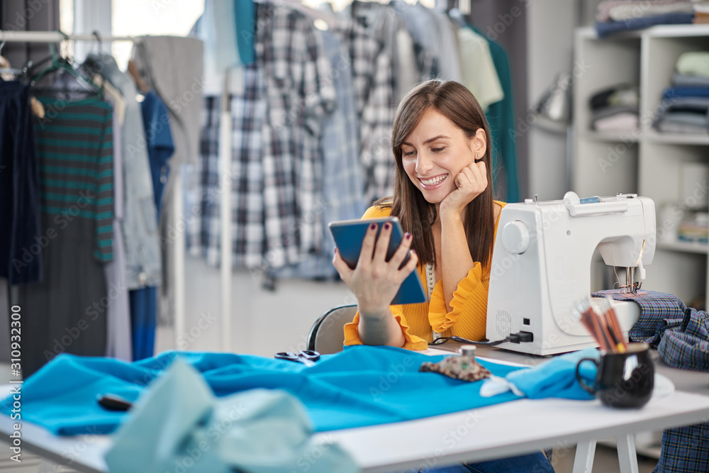 Obraz premium Smiling positive caucasian brunette sitting in her fashion studio and using tablet.