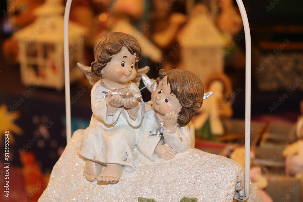 Statuette of two children angels in white sitting on a white cloud on a fuzzy background of other figurines.