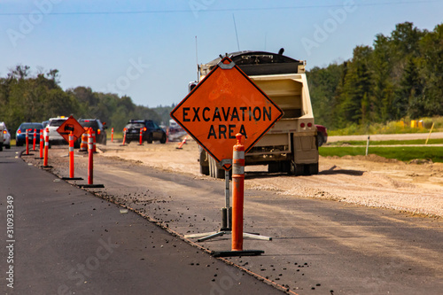 A close up shot of a bright orange warning sign saying excavation area is seen with traffic cones and construction vehicles in the background