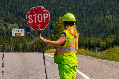 A close up and rear view of a female road construction worker holding a stop stick wearing high visibility safety clothes, roadworks traffic control