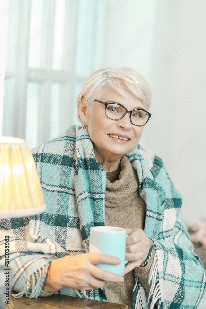 Senior Woman Wrapped In A Checkered Plaid Is Drinking Tea At Home Senior Woman Wrapped In A Checkered Plaid Is Drinking Tea At Home