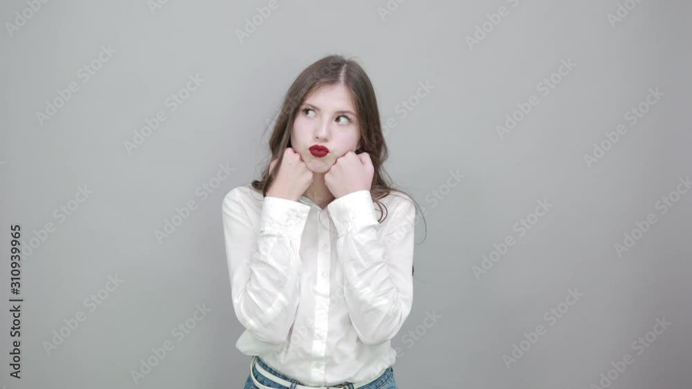 Attractive young woman in fashion white shirt isolated on gray background in studio holding hands over chin, looking pretty. People sincere emotions, lifestyle concept. Attractive young woman in fashion white shirt isolated on gray background in studio holding hands over chin, looking pretty. People sincere emotions, lifestyle concept.