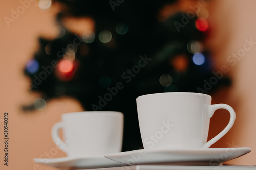 Two white color cups of coffee with Christmas tree in the background, focus is on the first one.