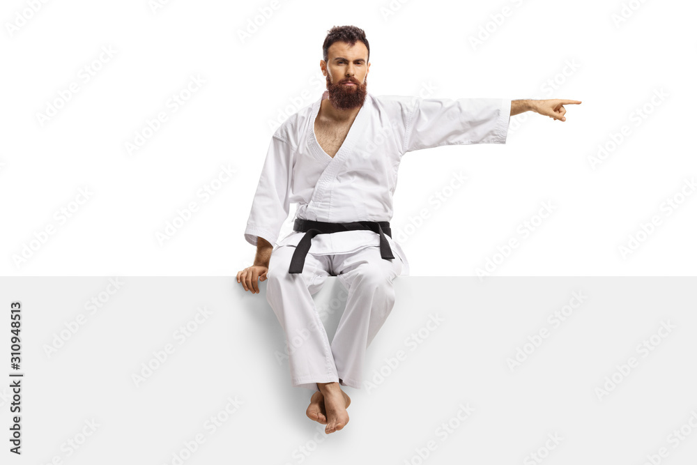Man in karate kimono with black belt sitting on a blank panel and pointing to one side Man in karate kimono with black belt sitting on a blank panel and pointing to one side