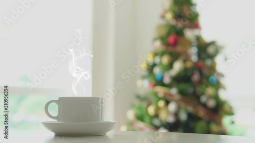 Close up white hot cup with smoke and blurred Christmas tree with blinking lights decoration.