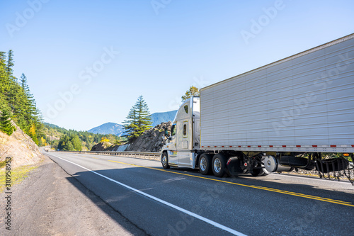 Big rig professional industrial grade semi truck transporting goods in refrigerator semi trailer running on the straight road with rocks and trees