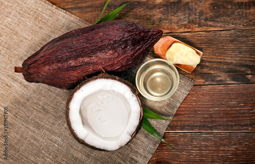 Half coconut with bottle of coconut oil, cocoa butter in a wooden scoop and cocoa pod with palm leaves on a wooden background.