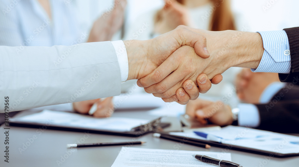 Business people shaking hands at meeting while theirs colleagues clapping and applauding. Group of unknown businessmen and women in modern white office. Success teamwork, partnership and handshake