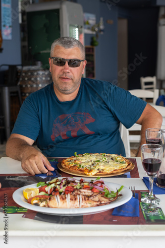 a man in a blue T-shirt and sunglasses sits at a table in a cafe in Europe. in front of him is a pizza and stuffed squid