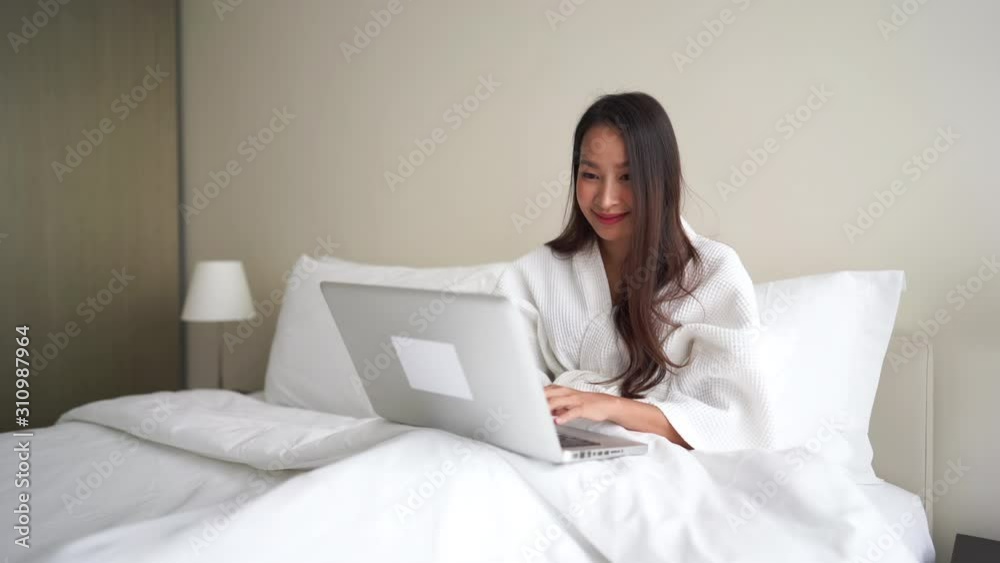 A young Asian woman dressed in a comfy white robe, sitting up in a resort hotel bed works on her laptop. A young Asian woman dressed in a comfy white robe, sitting up in a resort hotel bed works on her laptop.