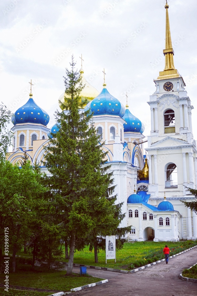Orthodox Church with blue and gold domes on a cloudy summer day, in front of the Church grow large fir trees