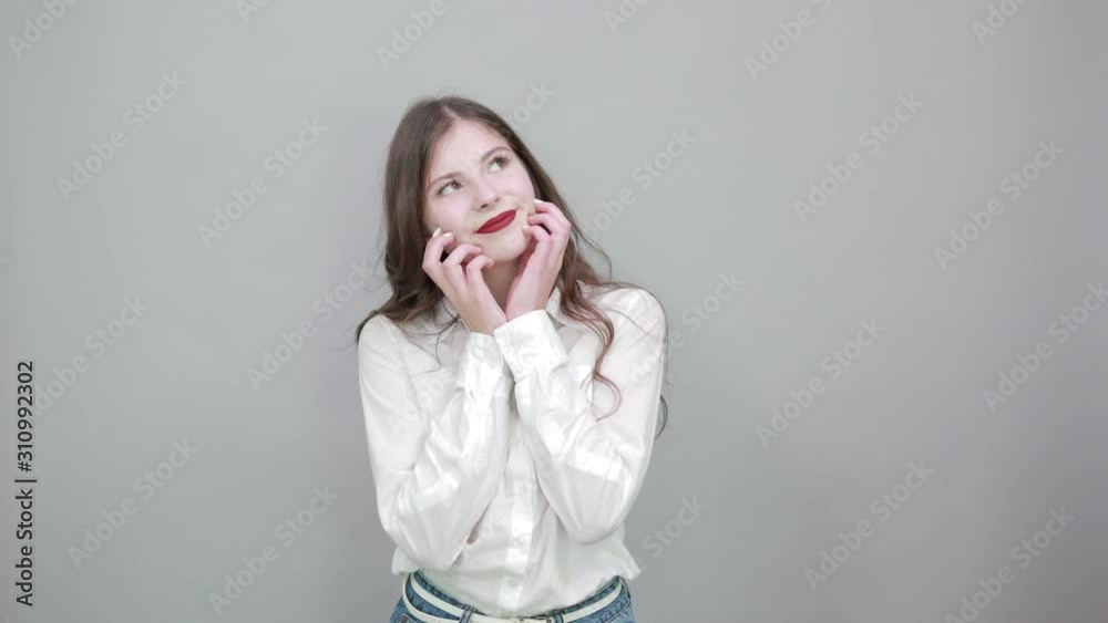 Attractive young brunette woman over isolated gray background wearing casual white shirt, looking up, holding hands over cheek, dreaming. People lifestyle concepte. Attractive young brunette woman over isolated gray background wearing casual white shirt, looking up, holding hands over cheek, dreaming. People lifestyle concepte.