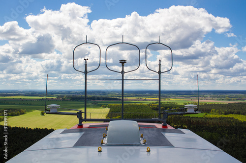 Measurement devices (cup anemometer, ultrasonic anemometer, wind vane) and aviation lights on the nacelle roof of a wind turbine with fields and cloudy sky in the background