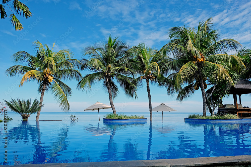 Luxury infinity swimming pool with blue water, umbrellas, palms and endless ocean view. Bali, Indonesia.