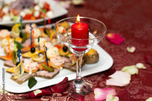 Fototapeta Naklejka Na Ścianę i Meble -  Beautifully laid table for the holiday. A candle in a glass goblet next to a plate of food appetizers on a red tablecloth in a restaurant.