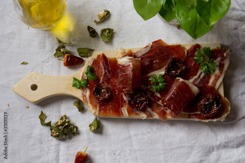  A cutting board with Traditional tapas with spanish cured meat, sun dried tomato , parsley, dry red and green pepper, green leaves on rustic lint tablecloth