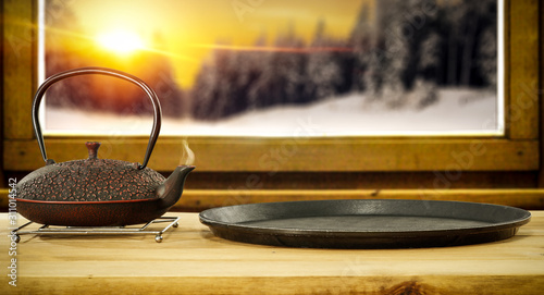 Wooden window sill with free space for your products. Board of kettle with hot drink of tea, coffee or mulled.Cold winter day and landscape of forest and orange sky with sunset time. Copy space. 