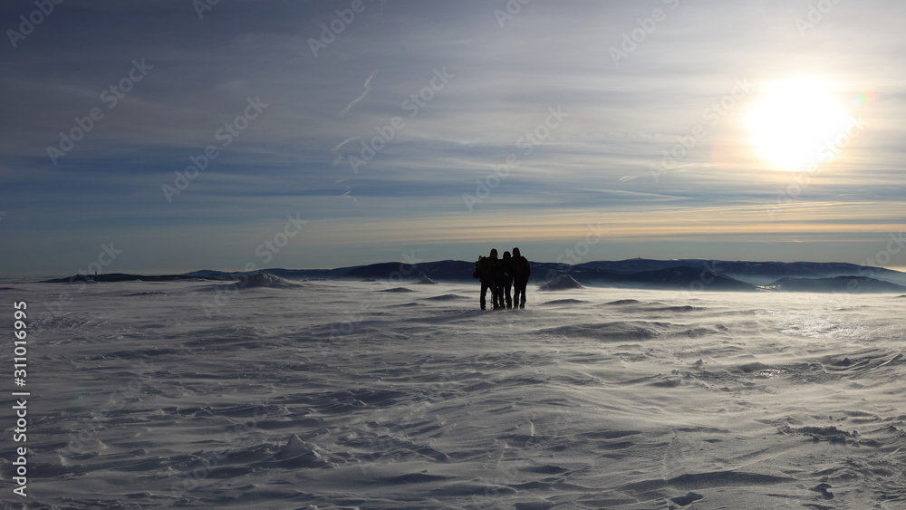 Silhouettes of three people at the top of snow covered mountain at the end of a day during sunset.