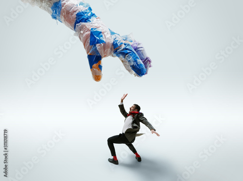 Fototapeta Naklejka Na Ścianę i Meble -  Young caucasian man avoiding big plastic hand on white studio background. Male model in office attire fighting in motion and action. Looks angry, scared, pressured. Eco problems, pollution, recycling.