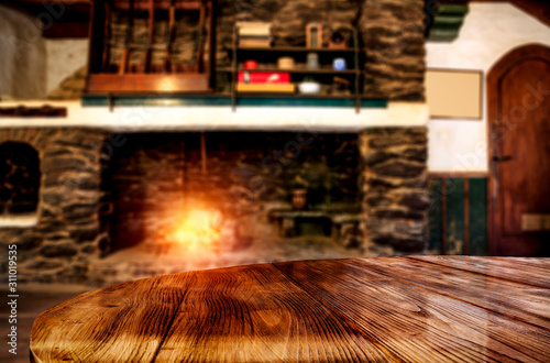 Wooden brown table top with space for your product.Interior of an old small house in the mountains with a fireplace.Flame with an orange color of burning wood in the hearth.Winter time and copy space.