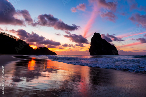 Fototapeta Naklejka Na Ścianę i Meble -  Stunning colorful sunset in Cacimba do Padre beach in Fernando de Noronha, Brazil with water reflection. Several clouds turned pink forming colorful rays in the sky and in the sand.