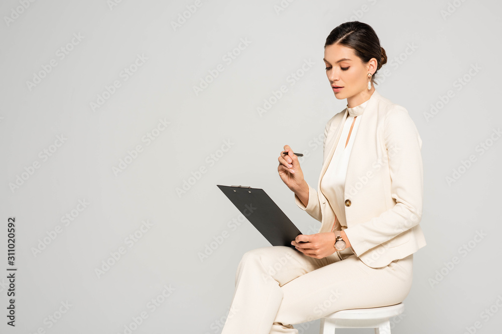 elegant attractive businesswoman in white suit sitting on stool with pen and clipboard, isolated on grey elegant attractive businesswoman in white suit sitting on stool with pen and clipboard, isolated on grey